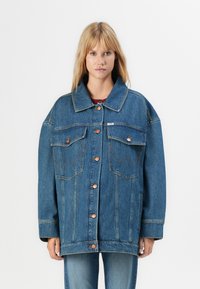 Woman wearing oversized blue denim jacket with button closure and flap chest pockets, paired with blue jeans against a plain white background.