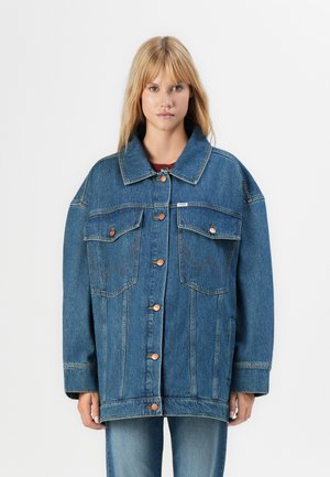 Donna che indossa una giacca di denim blu oversize con chiusura a bottoni e tasche a pattina sul petto, abbinata a jeans blu, su uno sfondo bianco semplice.