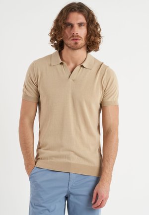 Uomo con capelli ricci che indossa una polo beige a maniche corte lavorata a maglia e pantaloni azzurri, in piedi davanti a uno sfondo bianco semplice.