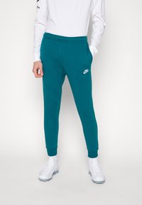 Man som bär teal Nike sweatpants, vit långärmad skjorta, vita strumpor och vita sneakers, som står mot en enkel bakgrund.
