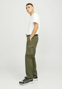 Giovane uomo di profilo che indossa una maglietta bianca, pantaloni cargo verde oliva con tasca e sneaker nero-grigie su sfondo semplice.