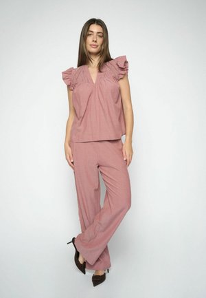 Woman standing wearing mauve ruffled short-sleeve top and wide-leg pants with black pointed high heels against white background.