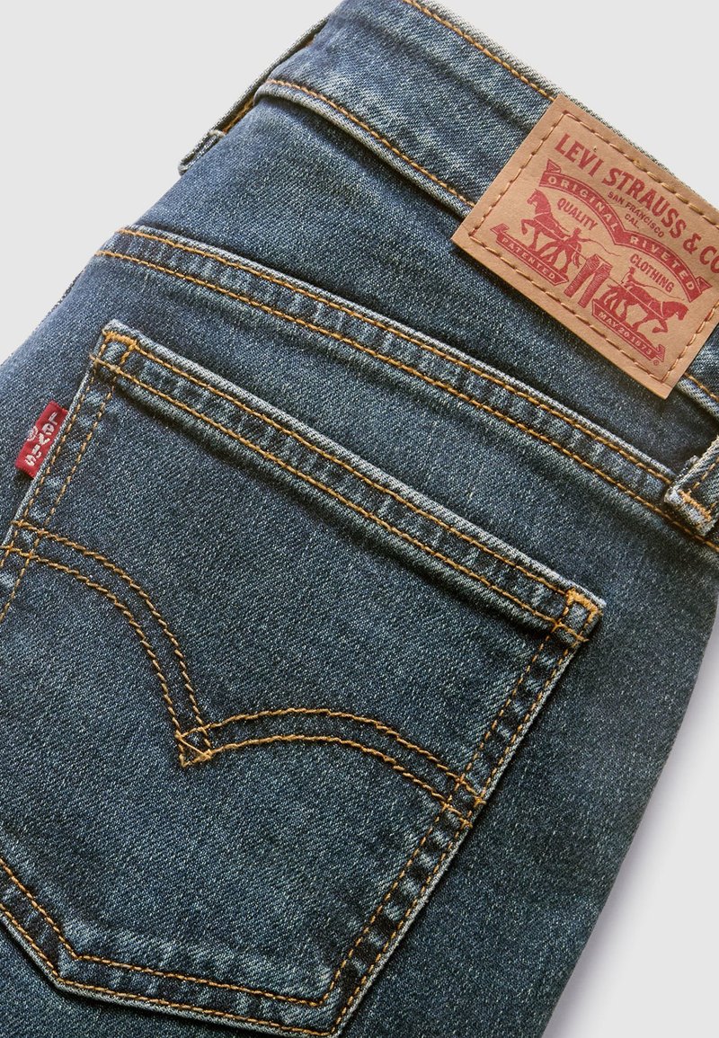 Dark blue denim jeans featuring a leather patch with embossed branding, orange stitching, and a signature back pocket design.