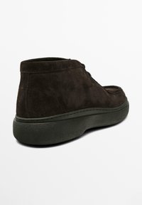 Braune Wildleder-Stiefeletten mit abgerundeter Spitze, schwarzen Schnürsenkeln und einer dicken grünen Gummisohle. Sie zeichnen sich durch eine glatte Textur und zurückhaltende Design-Details aus.