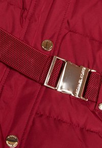 Viininpunainen untuvatakkikangas, jossa on sileä tekstuuri, leveä kudottu vyö ja kiiltävä kultaisen sävyinen solki, johon on kaiverrettu "MICHAEL KORS".