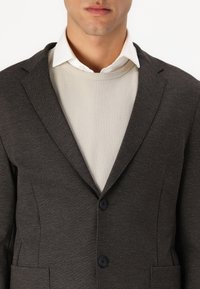 Donkergrijze blazer met een getailleerde pasvorm, twee knopen, geknipte revers en een fijne textuur, over een crèmekleurige ribbeltrui.