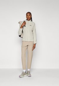 Veste en polaire gris clair avec un col montant, fermeture éclair, pantalon beige et baskets multicolores, avec un sac à dos porté sur une épaule.
