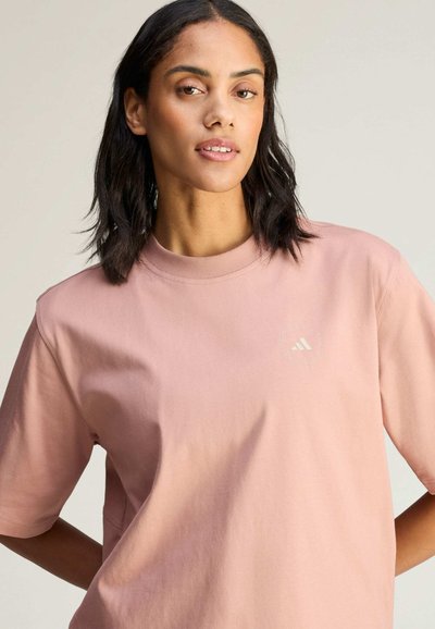 Woman with dark hair wears a loose-fitting pink Adidas t-shirt with a small logo on the left chest, standing against a plain background.