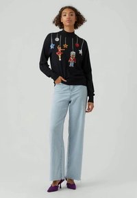 Frau in schwarzem Pullover mit hängender Nussknacker- und Balletttänzer-Dekoration, hellblauen weiten Jeans und lila Absätzen.