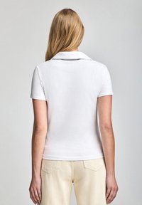 Camisa polo blanca con cuello clásico, mangas cortas y una textura suave. La camisa se combina con pantalones amarillos pálidos.