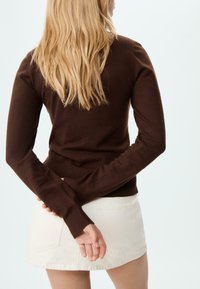 Braun gestrickter Pullover mit langen Ärmeln, gerippten Bündchen und Saum, kombiniert mit einem weißen Jeans-Minirock mit Gesäßtaschen. Klarer Hintergrund.