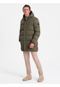 Olivengrøn pufjakke med hætte, lynlåslommer og et logo-patch på ærmet, parret med beige bukser og brune støvler.