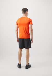 Īsas piedurknes oranžs sporta krekls, melni šorti un melnas kurpes. Iezīmē gludu tekstūru un pieguļošu dizainu, skatīts no aizmugures.