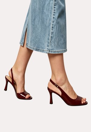 Personne portant une jupe en jean bleu mi-longue et des escarpins slingback en cuir verni bordeaux avec orteils ouverts et lanières à la cheville.