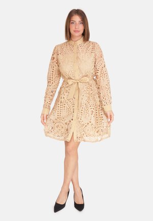 Beige kanten jurk met ingewikkelde uitsnijdingen, een knoopsluiting aan de voorkant, lange mouwen en een bijpassende riem om de taille. Zwarte hakken completeren de look.