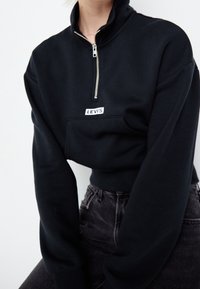 Persona che indossa un felpa nera Levi's con zip fino al collo e jeans neri a vita alta, tagliato sotto il mento su uno sfondo semplice.