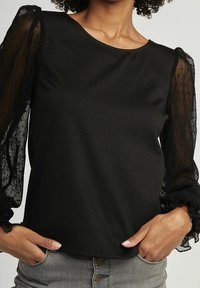 Top nero con maniche trasparenti e testurizzate, scollatura rotonda e vestibilità rilassata. Presenta polsini volant per un tocco di dettaglio in più.