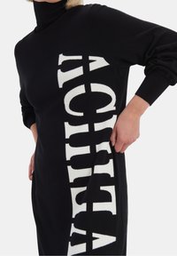 Svart stickad klänning med hög krage och överdimensionerad vit text på sidan. Strukturerad tyg med ribbade manschetter.