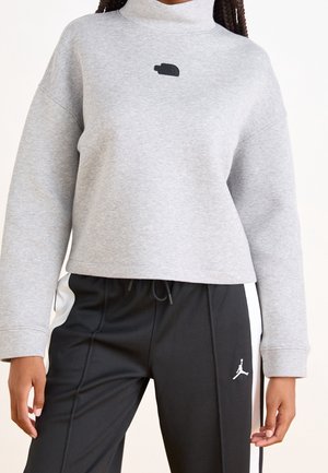 Person iført lysegrå cropped sweatshirt med høj krave og sorte atletikbukser med hvide sidestriber og et lille logo.