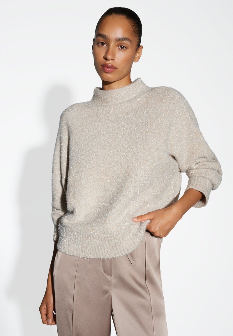 Pull en tricot beige avec un col montant, coupe légèrement ample et poignets côtelés, associé à un pantalon en tissu lisse couleur beige clair.
