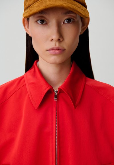 Giacca rossa con zip e colletto appuntito, caratterizzata da una texture liscia e una cerniera argento. Un berretto in velluto a coste color camoscio completa il look.