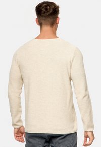 Pull en tricot beige avec une finition texturée, manches longues et col rond. Présente des fentes latérales au niveau de l'ourlet pour une aisance de mouvement.