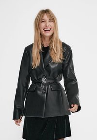 Blazer en cuir noir avec une fermeture à un seul bouton et deux poches avant. Dispose d'une large ceinture avec un accent décoratif en argent.
