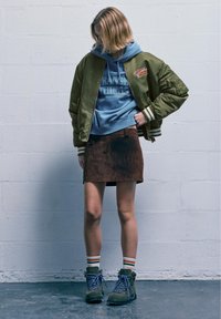 Oliivivihreä pomppari, vaaleansininen huppari, jossa on "Stranger Things" -logo, ruskea mokkahame, vihreät vaelluskengät sinisillä nauhoilla, raidalliset sukat.