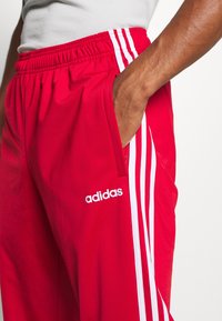 Czerwone spodnie dresowe z białymi trzema paskami i elastycznym pasem. Posiadają kieszenie i logo Adidas na udzie.