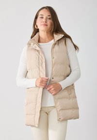 Gilet matelassé beige avec fermeture éclair et boutons-pression, col montant, poches latérales et texture matelassée, porté sur une chemise blanche à manches longues.