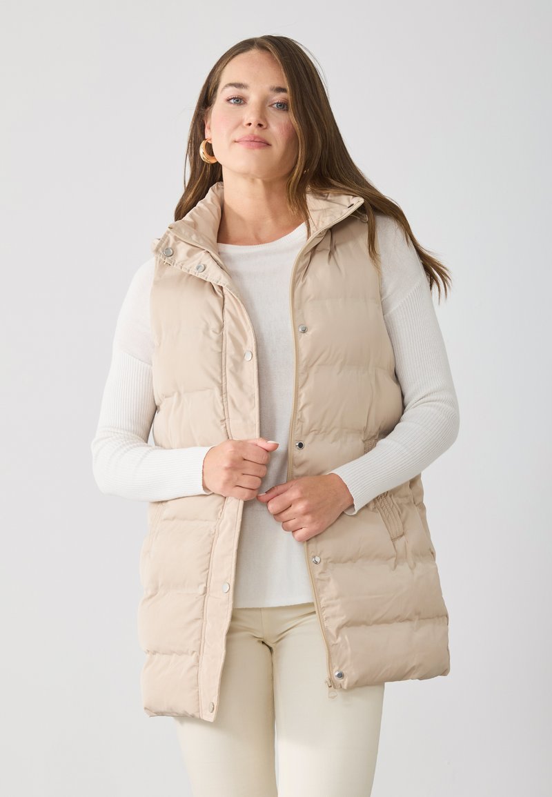 Gilet matelassé beige avec fermeture éclair et boutons-pression, col montant, poches latérales et texture matelassée, porté sur une chemise blanche à manches longues.