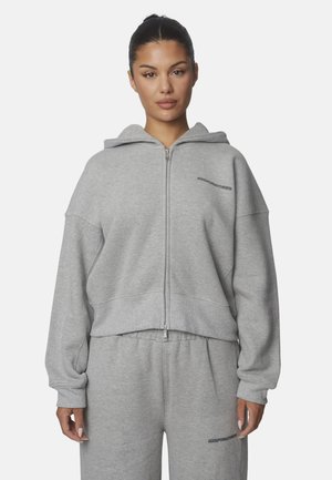 Vrouw met donker haar in een knot draagt een lichtgrijze zip-up hoodie en bijpassende joggingbroek met kleine tekstlogo's op borst en dij.
