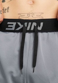 Šedé športové šortky s čiernym elastickým pásom, ktorý obsahuje šedé logo "NIKE", doplnené čiernymi šnúrkami a textúrovanou látkou.