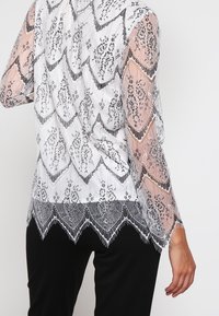 Top in pizzo con orlo a onde, motivo floreale in bianco e nero, maniche lunghe trasparenti e dettagli in frange intricate nella parte inferiore.