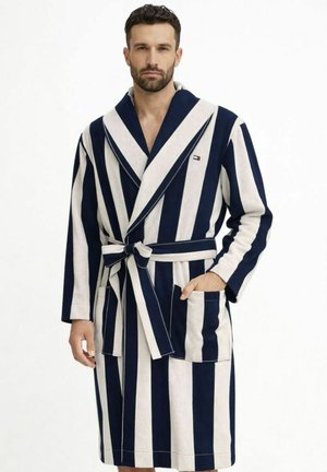 Uomo che indossa un accappatoio a righe verticali blu navy e bianche, con cintura annodata in vita e una mano in tasca.