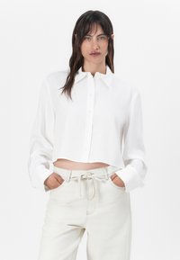 Cropped white button-up shirt made of smooth fabric, featuring a classic collar and long sleeves, paired with light-colored high-waisted pants.