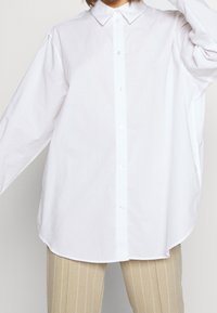 Chemise blanche à boutons avec col pointu, manches longues et coupe décontractée. Tissu lisse et léger avec une texture unie.