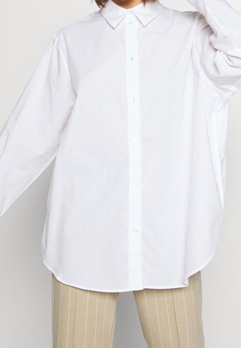 Chemise blanche à boutons avec col pointu, manches longues et coupe décontractée. Tissu lisse et léger avec une texture unie.