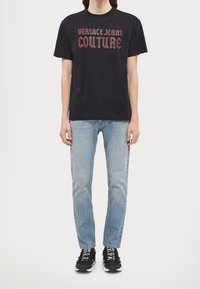 Černé bavlněné tričko s červeným nápisem "VERSACE JEANS COUTURE", spojené se světle modrými džínami a černými teniskami s bílými akcenty.