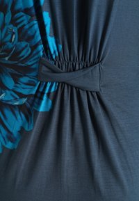 Tummet blår kläde med blommönster i aqua, med en samlad midja med knuten detalj. Mjuk textur, lätt material.