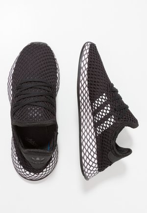 Zapatillas Adidas negras con patrón de malla blanca en la suela y la parte superior, con tres franjas blancas en los lados y cordones negros.