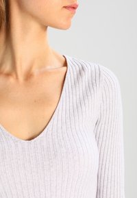 Haut gris clair en maille côtelée avec un profond décolleté en V, une silhouette ajustée et un tissu texturé, mettant en valeur les détails du cou et des épaules.