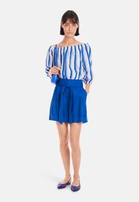 Blaue schulterfreie Bluse mit weißen Streifen, kombiniert mit hochtaillierten, gefalteten blauen Shorts und passenden blauen Ballerinas. Eine schlichte Handtasche ist ebenfalls enthalten.