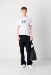 White cotton t-shirt with colorful "Calvin Klein" logo, paired with dark denim jeans and a black tote bag, worn with white sneakers.