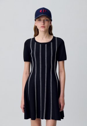 Donna che indossa un vestito nero a maniche corte con righe verticali bianche e un berretto blu navy con un logo rosso, in piedi davanti a uno sfondo semplice.