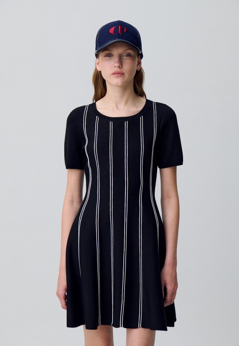 Mujer con un vestido negro de manga corta y rayas blancas verticales, y una gorra azul marino con un logo rojo, de pie contra un fondo sencillo.