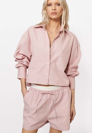 Blondynka ubrana w luźną, jasnoróżową koszulę w cienkie paski z długim rękawem i pasujące szorty ze sznurkiem, stojąca z rękami w kieszeniach.