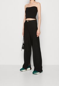 Woman wearing black strapless crop top, high-waisted wide-leg black trousers with front slits, green and white sneakers, holding small black bag.