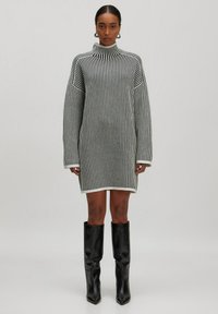 Robe en tricot à rayures verticales noires et blanches, avec un col montant, des manches oversize et une silhouette droite. Portée avec des bottes noires jusqu'aux genoux.