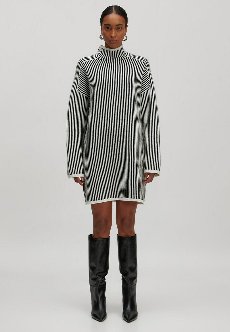 Robe en tricot à rayures verticales noires et blanches, avec un col montant, des manches oversize et une silhouette droite. Portée avec des bottes noires jusqu'aux genoux.